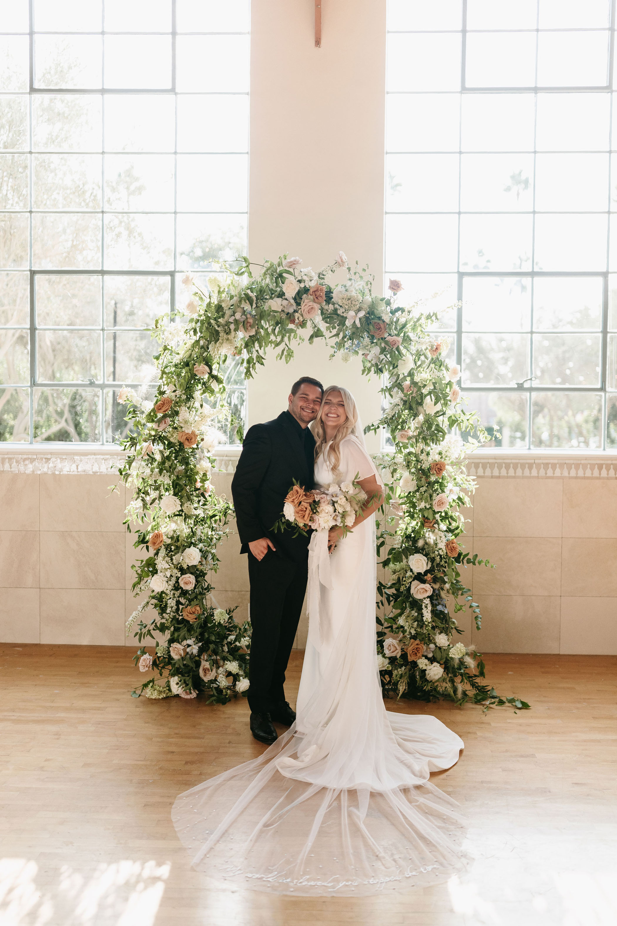 bride and groom smiling under floral ceremony arch in windowed indoor venue