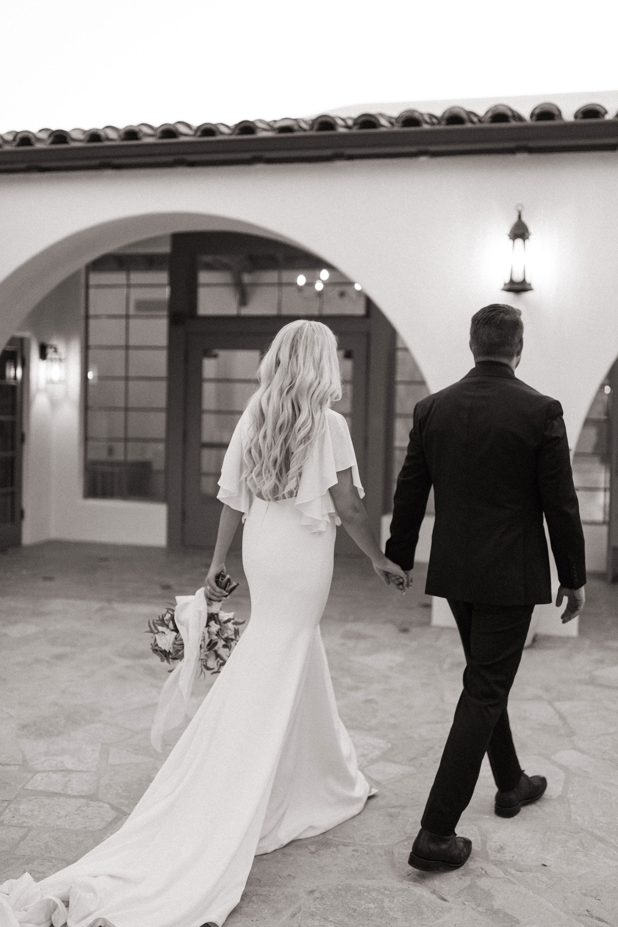 black and white photo of bride and groom holding hands walking towards their wedding reception