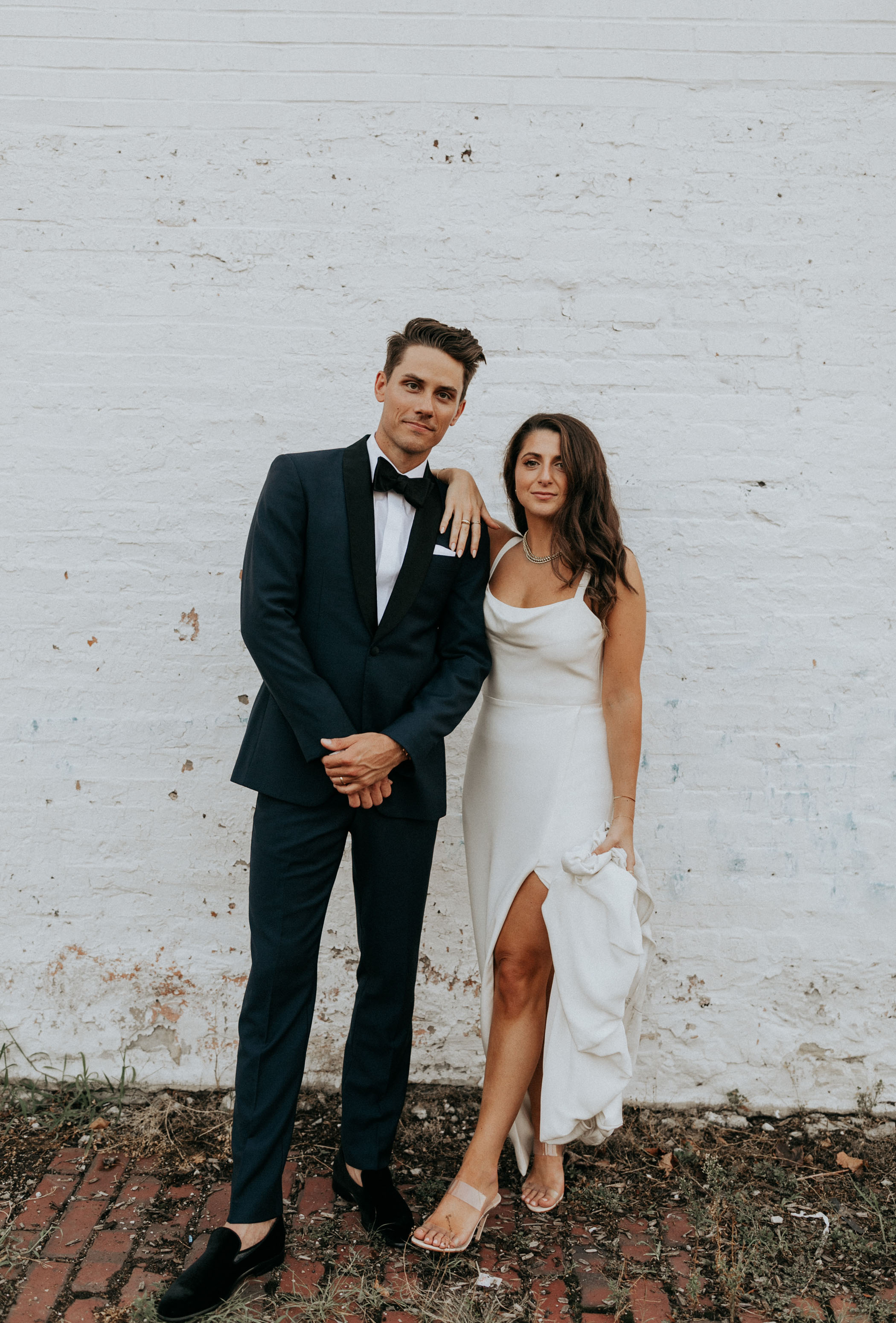 bride and groom posing against white brick wall