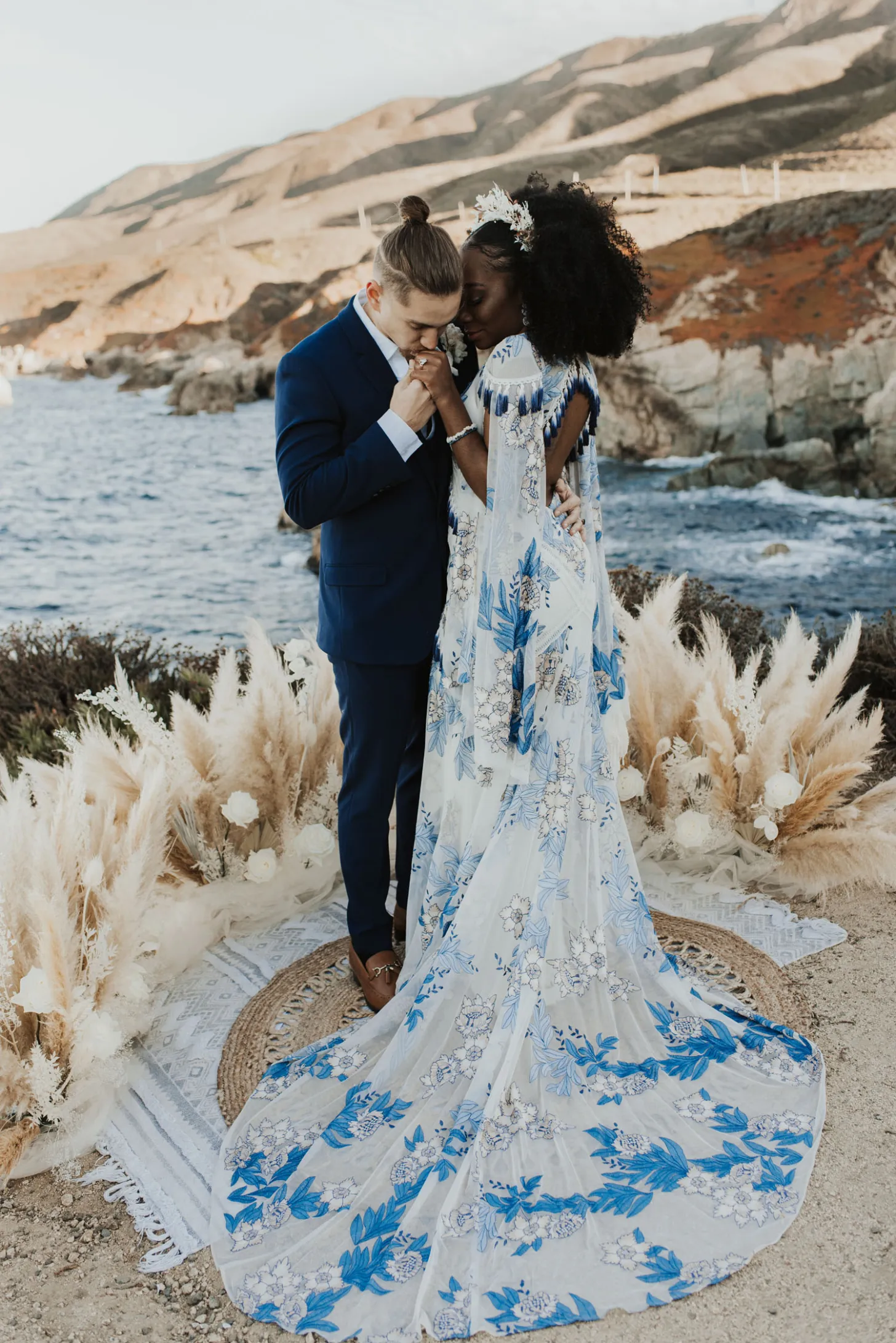 groom kissing bride's hand as they stand at their ceremony site on a cliff by the ocean during their wedding planned in Big Sur at sunset