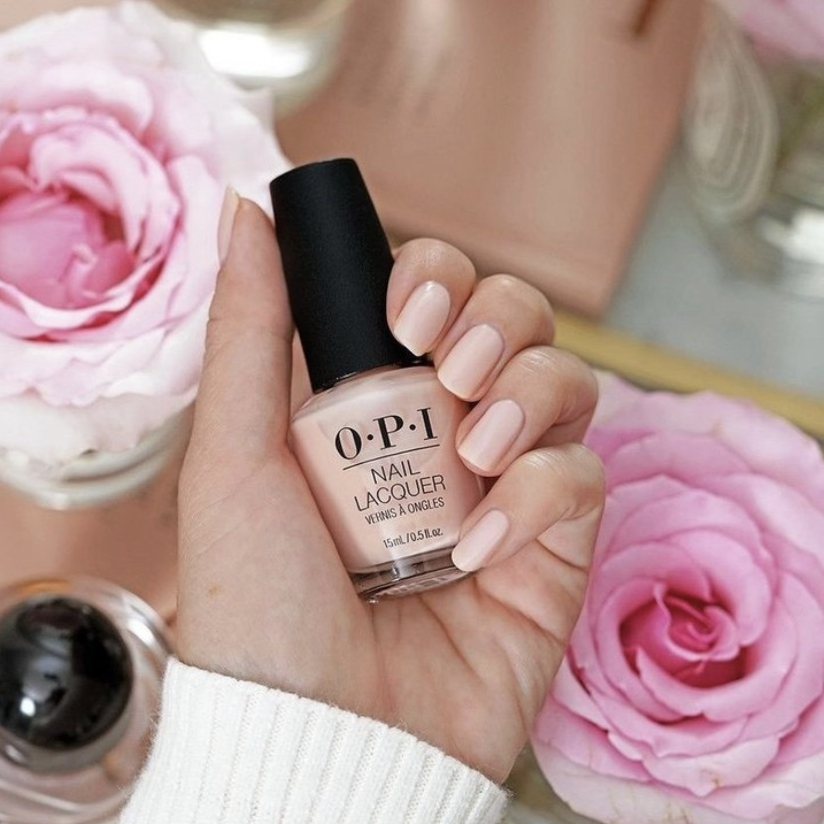 a woman with a nude pink manicure holding a bottle of matching nude OPI affordable drugstore nail polish in front of a floral background