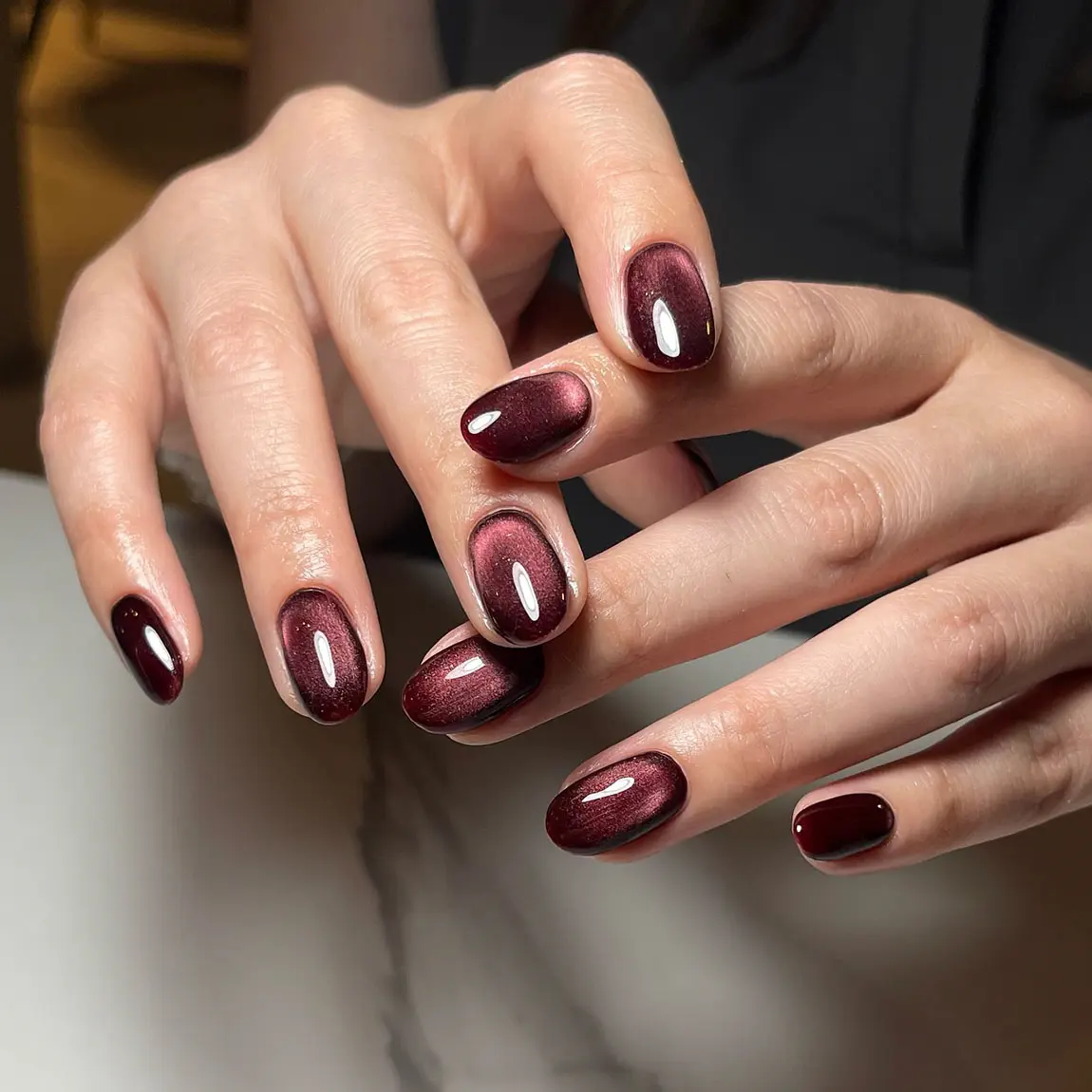 Short round burgundy cat eye nails with a copper shimmer band on a white marble table